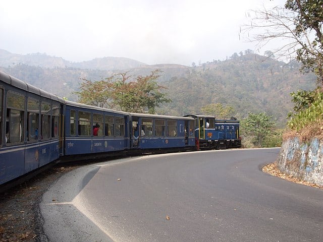 Puffing through misty Himalayan slopes, the Darjeeling ‘Toy Train’ offers a nostalgic ride past tea gardens, hill towns, and breathtaking mountain views, chugging along narrow tracks that cling to the hillsides on one of India’s most iconic rail journeys. Courtesy: Wikimedia